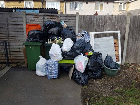 A collection of various black, white, and patterned rubbish bags, some tied and others open, are piled on a green bench and on the ground in an outdoor area near a wooden fence. Behind the bags, there are two large green wheelie bins, one with an orange lid and the other without a lid, along with a green plastic plant pot on the right side. An upturned wooden bed frame or pallet is leaning against the fence on the right. The scene appears to be a designated area for waste or rubbish collection, possibly on a residential driveway or alleyway, with buildings visible in the background. The ground is paved with asphalt, and there is some exposed soil and minimal vegetation near the fence. The lighting is natural, indicating an overcast day. This image represents waste collection and rubbish removal services, potentially offered by Commercial Waste Removal Crystal Palace, serving the local postcode area.