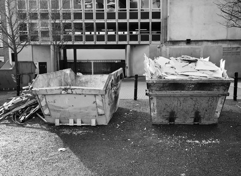 The image shows two large industrial skips positioned on a paved surface in front of a modern concrete building, likely situated in Crystal Palace. The left skip, which appears to be made of metal and painted in a light color, is filled with broken pieces of plasterboard and construction debris with rough, uneven edges and a dusty surface. The right skip, constructed from dark weathered metal, contains a sizable pile of crumpled white paper and cardboard waste, some of which slightly spills over the top edges. The surrounding environment includes a concrete pavement, a background of a multi-story building with a grid-patterned facade, and a few small bollards separating the waste area from the building. On the left side, a small tree with leafless branches indicates the photo was taken in winter or early spring. This scene relates to commercial waste removal services, such as those offered by Commercial Waste Removal Crystal Palace, for disposing of construction and paper waste efficiently in the local area between the town and postcode area.