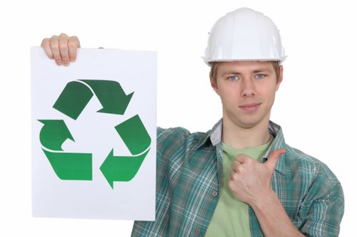 A young man wearing a white safety helmet and casual green plaid shirt is standing outdoors on a paved area, holding a white sign with a large green recycling symbol printed on it. His expression is neutral, and he is giving a thumbs-up gesture with his right hand, which is slightly raised near his chest. The background appears to be a bright, clear day with a plain, light-colored backdrop, emphasizing the focus on the man and the recycling sign. This image relates to environmental awareness and waste management, aligning with services offered by Commercial Waste Removal Crystal Palace in the local postcode area, suggesting a focus on responsible rubbish disposal and recycling practices in the Crystal Palace region.