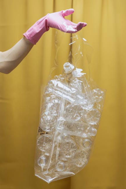 A person wearing a pink rubber glove is holding a large, transparent plastic bag filled with clear, crushed PET bottles and plastic waste, against a yellow curtain background. The plastic bottles insi