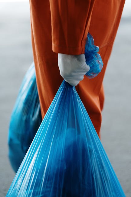 A person wearing a pink rubber glove is holding a large, transparent plastic bag filled with clear, crushed PET bottles and plastic waste, against a yellow curtain background. The plastic bottles insi
