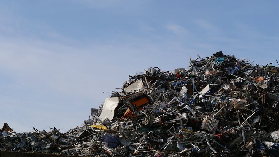 A large piles of assorted metal and electronic waste materials, including broken appliances, discarded machinery, and tangled cables, are stacked outdoors against a clear blue sky. The debris appears 