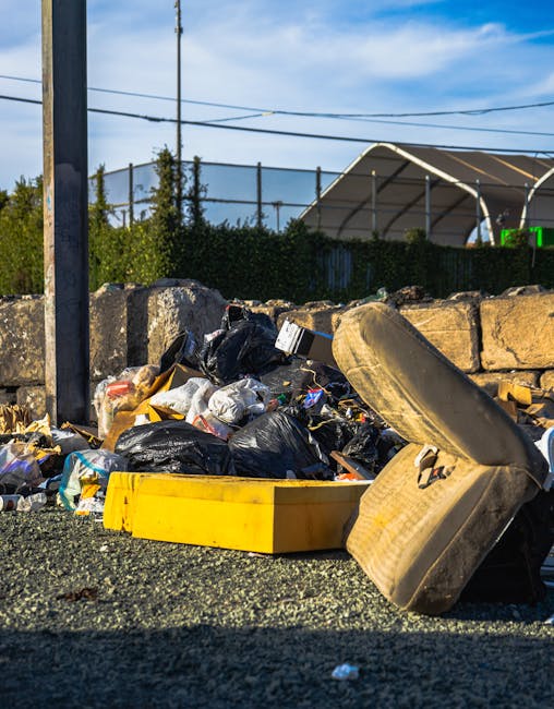 A large piles of assorted metal and electronic waste materials, including broken appliances, discarded machinery, and tangled cables, are stacked outdoors against a clear blue sky. The debris appears 