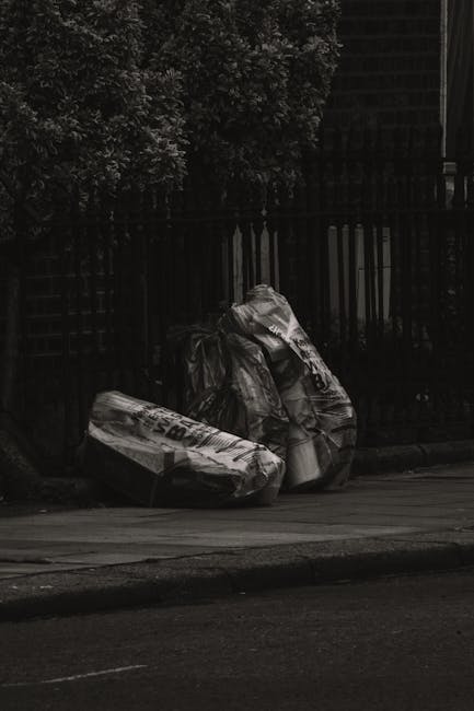 Two large black plastic bags filled with waste material are placed on the pavement next to a black metal fence, with one of the bags appearing crumpled and partially open. Behind the fence, dense foli