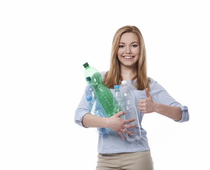 A young woman with shoulder-length red hair smiling and giving a thumbs-up gesture while holding several clear plastic bottles, including one green-tinted and two transparent with blue caps, against a plain white background. She is dressed in a light blue shirt with rolled-up sleeves and beige trousers. The bottles appear to be empty and are held close to her chest with both arms. The image is styled to convey a positive attitude towards recycling or waste management, relevant to rubbish removal services offered by companies like Commercial Waste Removal Crystal Palace. The plain white background provides a neutral setting that emphasizes the bottles and the woman's expression. The scene is brightly lit with no visible environmental context or background details, focusing solely on the subject and the bottles, suitable for illustrating waste reduction or rubbish disposal themes in a professional, straightforward manner.