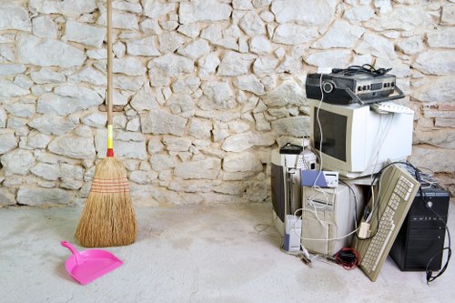 The image depicts a clear concrete floor area adjacent to a textured stone wall, with a wooden broom leaning against the wall on the left side. On the floor, there is a small bright pink dustpan positioned in front of the broom. To the right, a collection of discarded electronic waste and household appliances is stacked, including an old desktop computer monitor, a tower CPU, a keyboard, a pair of speakers, and a CRT television. Some cables are loosely draped over the devices. The setting appears to be an indoor or sheltered outdoor space, typical for a rubbish collection site, possibly in a residential or commercial area of Crystal Palace. The scene showcases typical items intended for disposal, with a focus on electronic waste and general refuse, aligning with rubbish removal services offered by Commercial Waste Removal Crystal Palace. The environment is well-lit, highlighting the textures of the stone wall and the various materials of the discarded items, reflecting a typical scene of waste awaiting removal in a local setting.