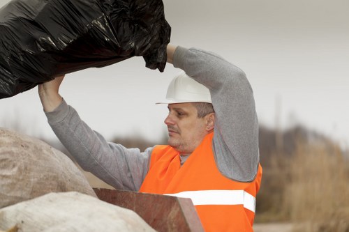 A male waste collection worker wearing a white safety helmet and an orange high-visibility vest with reflective stripes is outdoors, lifting a black plastic rubbish bag above a large pile of mixed waste materials. The waste pile contains various items, including brown and beige sacks and pieces of discarded material, situated on a paved or gravel surface. The background features an overcast sky and blurred natural landscape elements, indicating a waste removal operation possibly near a commercial or residential area in or around Crystal Palace, south London. The worker is focused on the task of lifting or sorting rubbish as part of a commercial waste removal service conducted by Commercial Waste Removal Crystal Palace, with the environment suggesting a typical rubbish collection or disposal scene associated with the company's services in the local area.